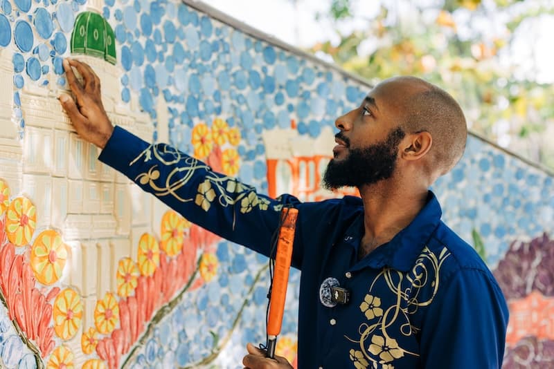 A man with a white cane reaching up to touch a raised green ceramic dome on the mosaic wall