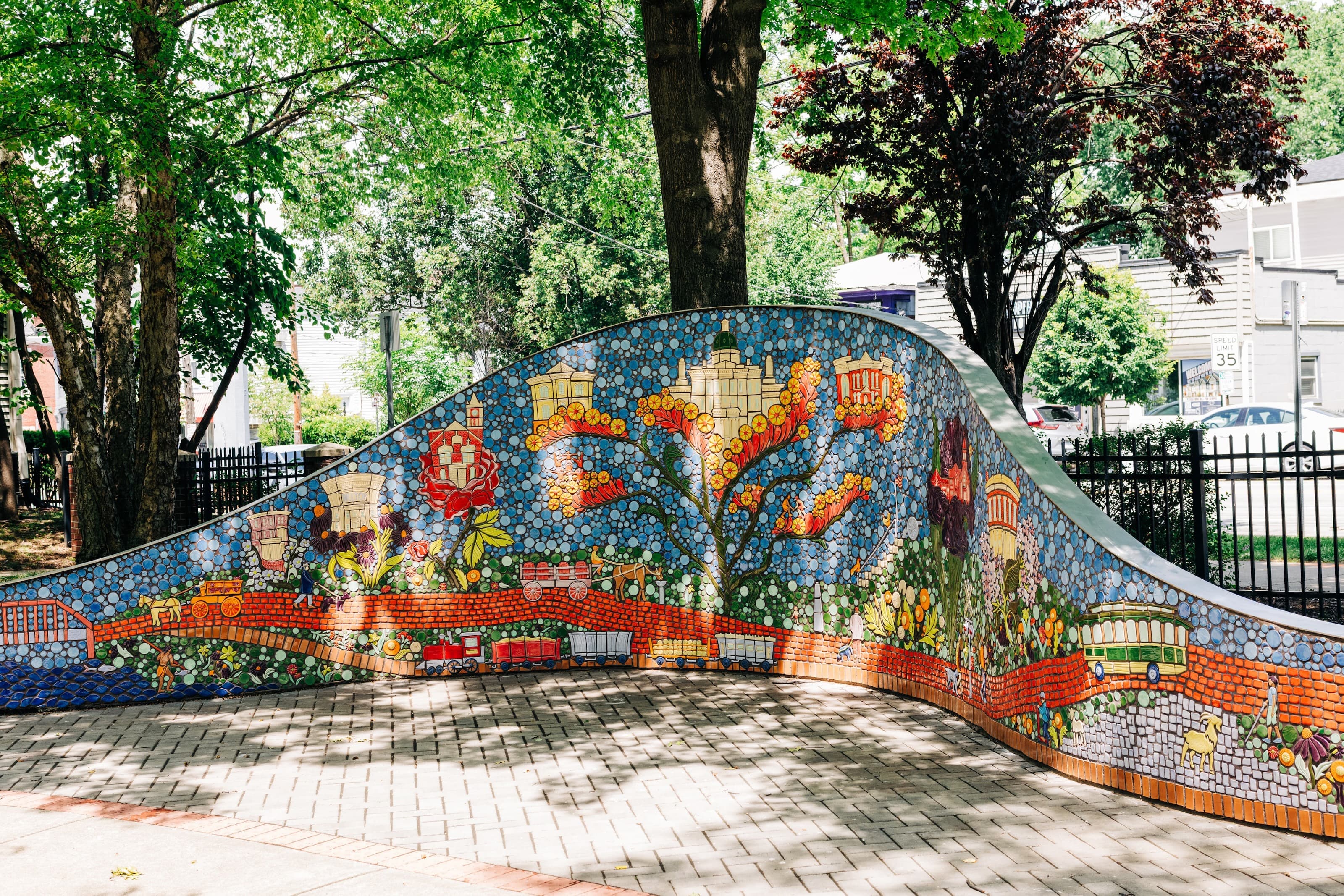 The finished wave-shaped ceramic mosaic wall curving through a tree-shaded brick plaza, its tiles depicting stylized buildings, a blossoming tree, wagons, and flowers