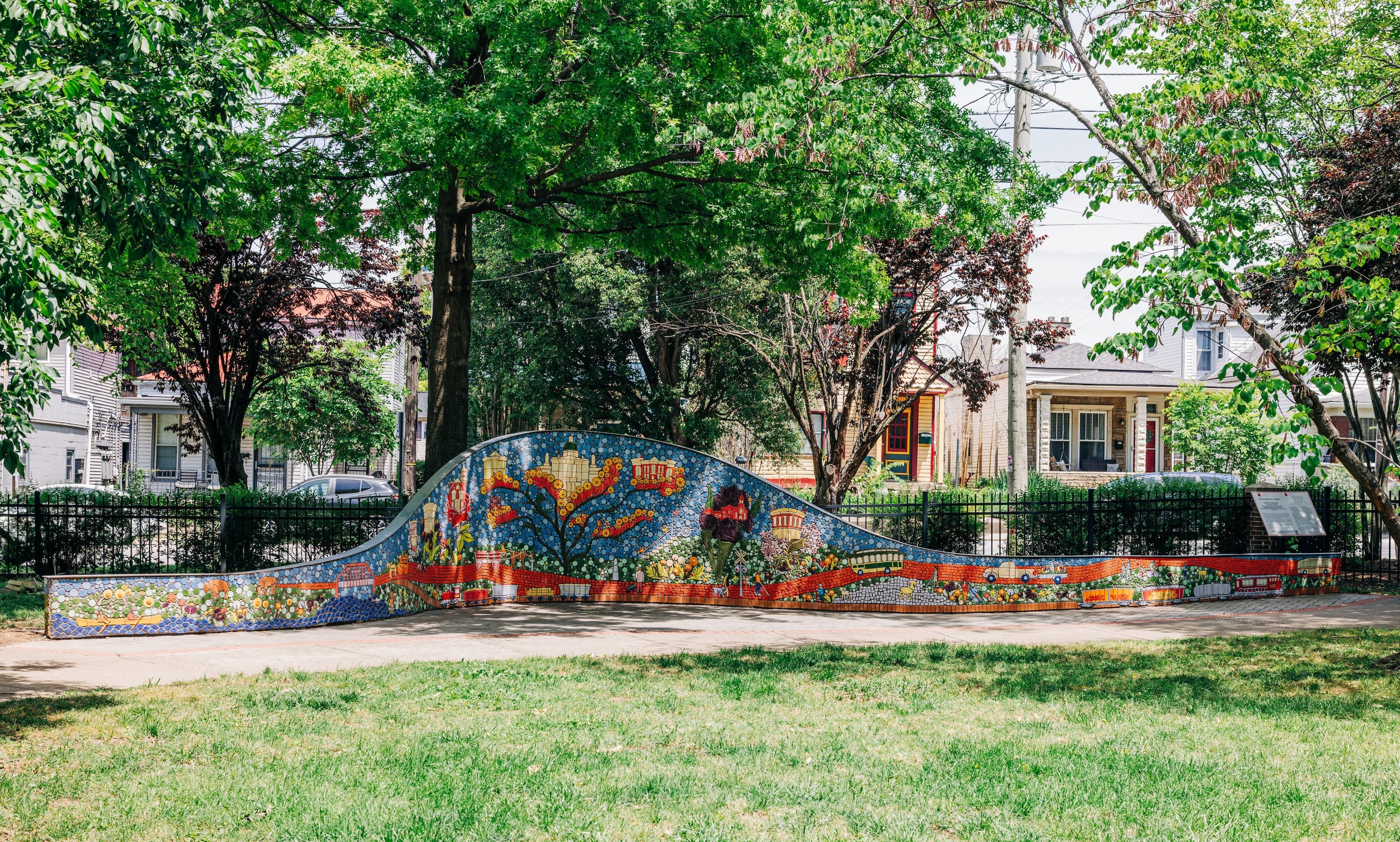 A wide view of the low, wave-shaped ceramic mosaic wall curving through a tree-lined Clifton park, with neighborhood houses visible beyond the fence behind it