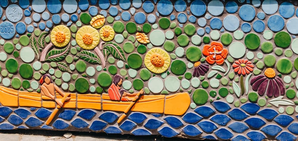 A mosaic panel of two people paddling a canoe across stylized blue water, beneath sunflowers and coneflowers