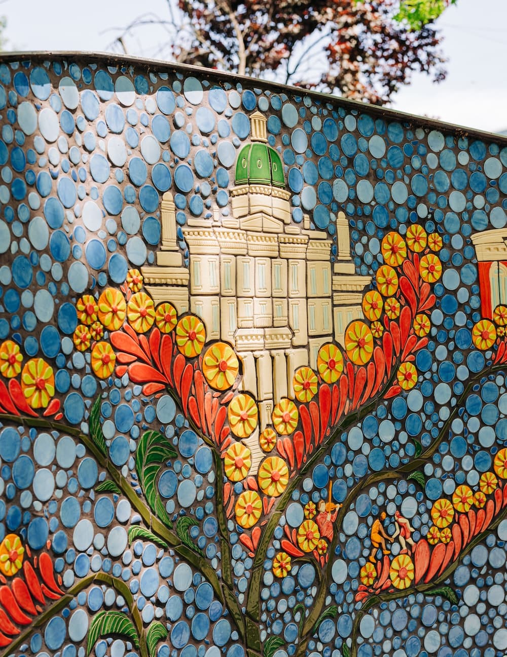 A mosaic panel of a domed capitol-style building framed by red petals and orange starburst flowers, with tiny wrestlers among the blooms
