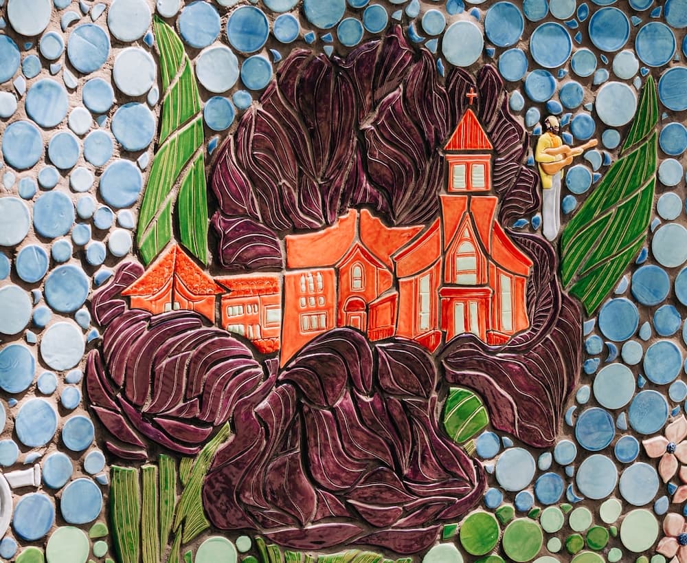 A mosaic panel of a large purple iris framing orange-glazed theater and church buildings, with a tiny figure playing guitar