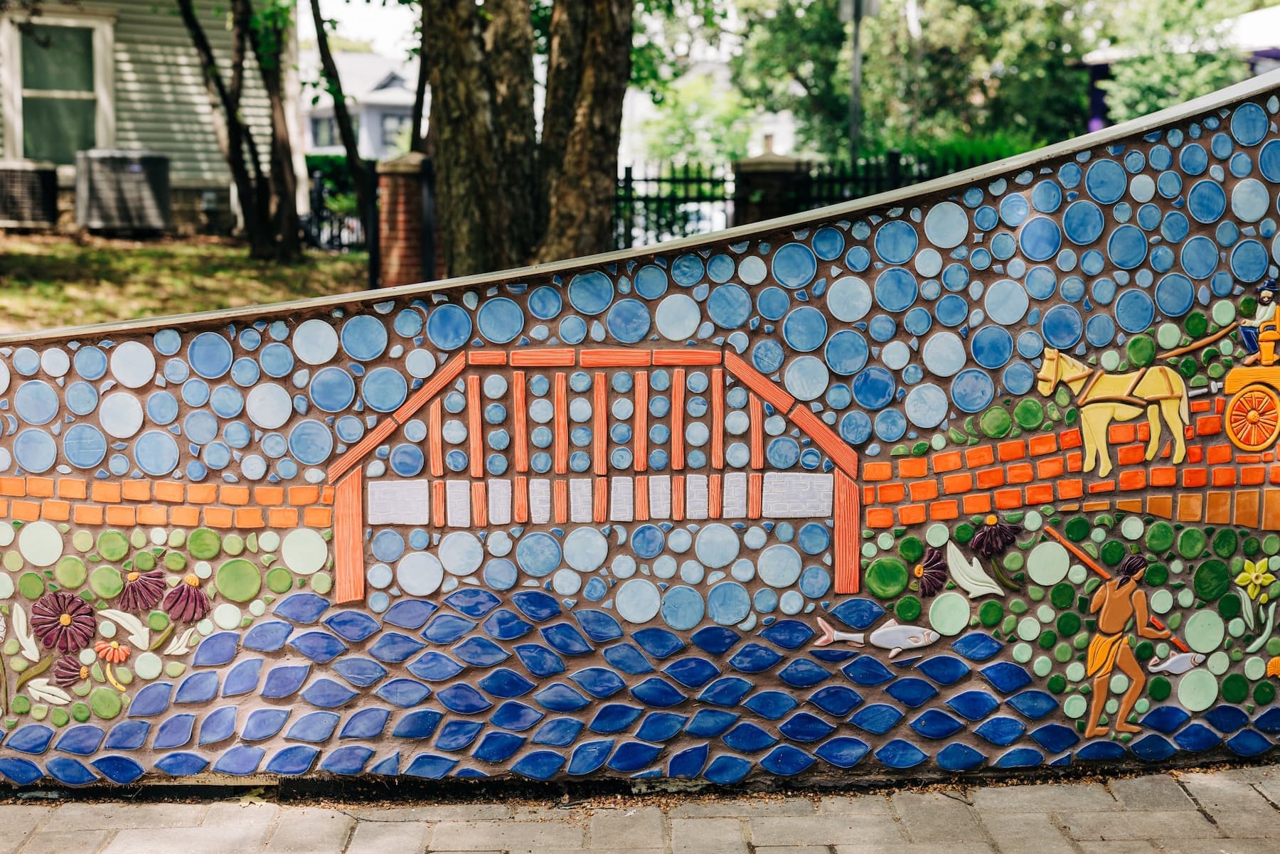 A mosaic panel detail of an orange covered bridge over Beargrass Creek, with a horse-drawn carriage on the brick path above and an indigenous fisher spearing fish in the blue water below