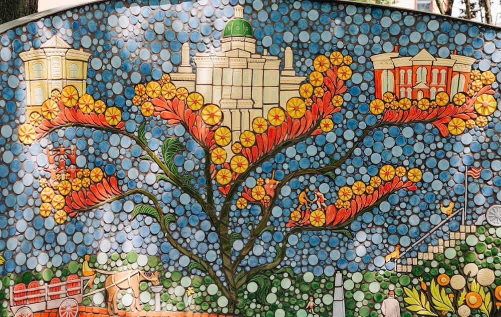 A mosaic panel of a branching tree bearing yellow blossoms and red petals, topped with ornate buildings above a horse-drawn wagon