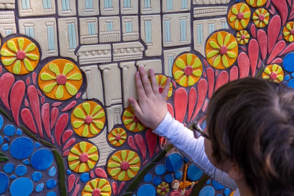 A child reaching up to touch the ceramic mosaic wall, a hand resting on the raised cream building tiles surrounded by orange and lime-green flowers